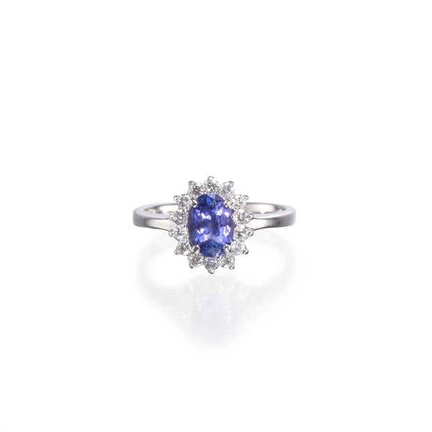 An oval-cut blue tanzanite ring set in a silver-colored halo with small clear accent stones, centered on a white background with a soft reflection.