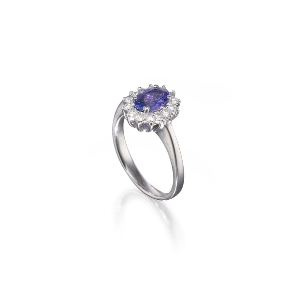 A white gold ring featuring a central oval blue tanzanite gemstone surrounded by a halo of small clear stones, set on a polished silver band against a white background.