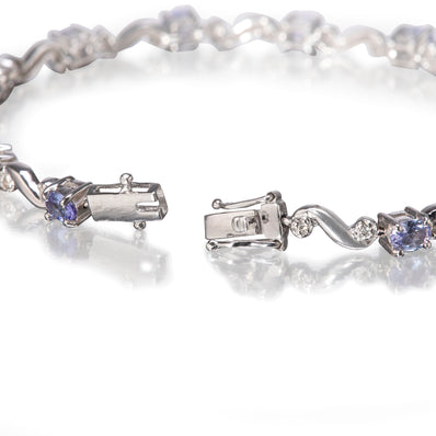A white gold tennis bracelet with round blue tanzanite stones and clear accent stones, shown in a close-up on a reflective white surface.