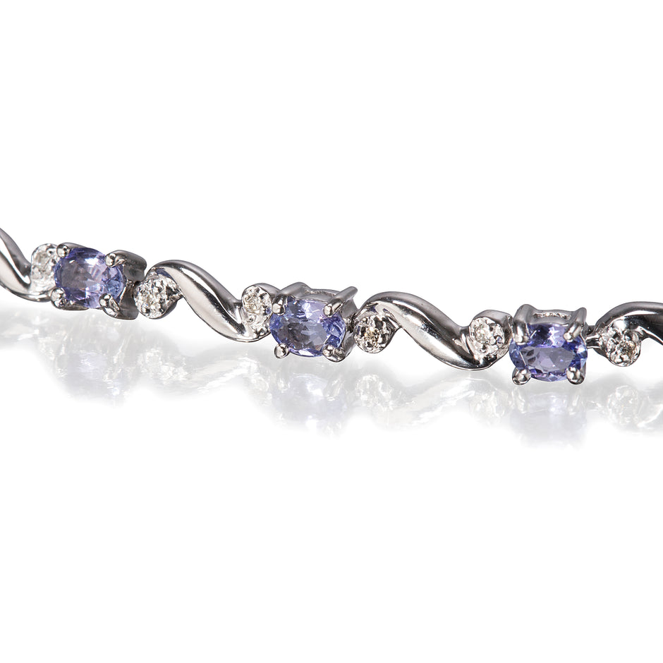 A close-up of a white gold tennis bracelet featuring oval tanzanite and round clear accent stones, set against a white background with a soft reflection.