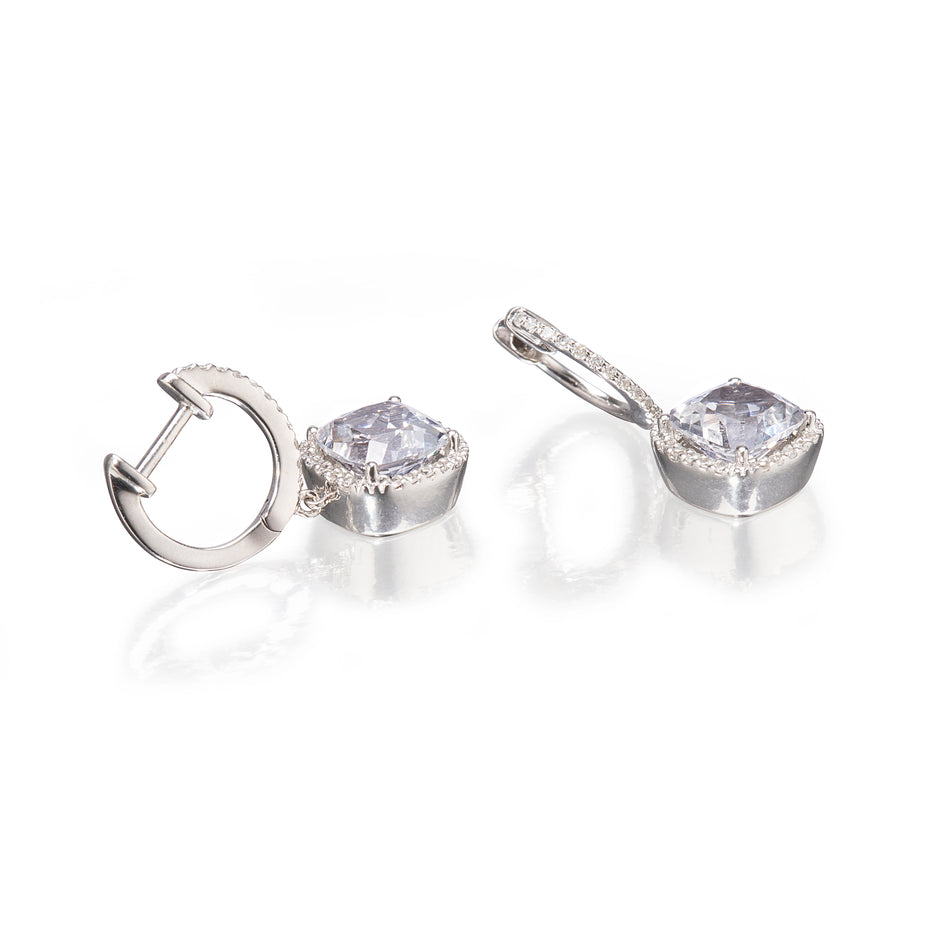 A pair of white gold huggie hoop earrings with cushion-cut clear gemstones and small accent stones, displayed on a reflective white surface.