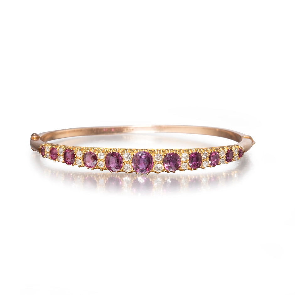 A rose gold bangle bracelet featuring a row of oval purple amethyst gemstones and small clear accent stones, shown on a clean white background with a soft reflection.