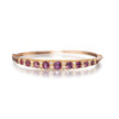 A rose gold bangle bracelet featuring a row of oval purple amethyst gemstones and small clear accent stones, shown on a clean white background with a soft reflection.