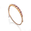 A gold hinged bangle bracelet featuring a row of oval pinkish-red gemstones and small clear stones, shown on a clean white background.