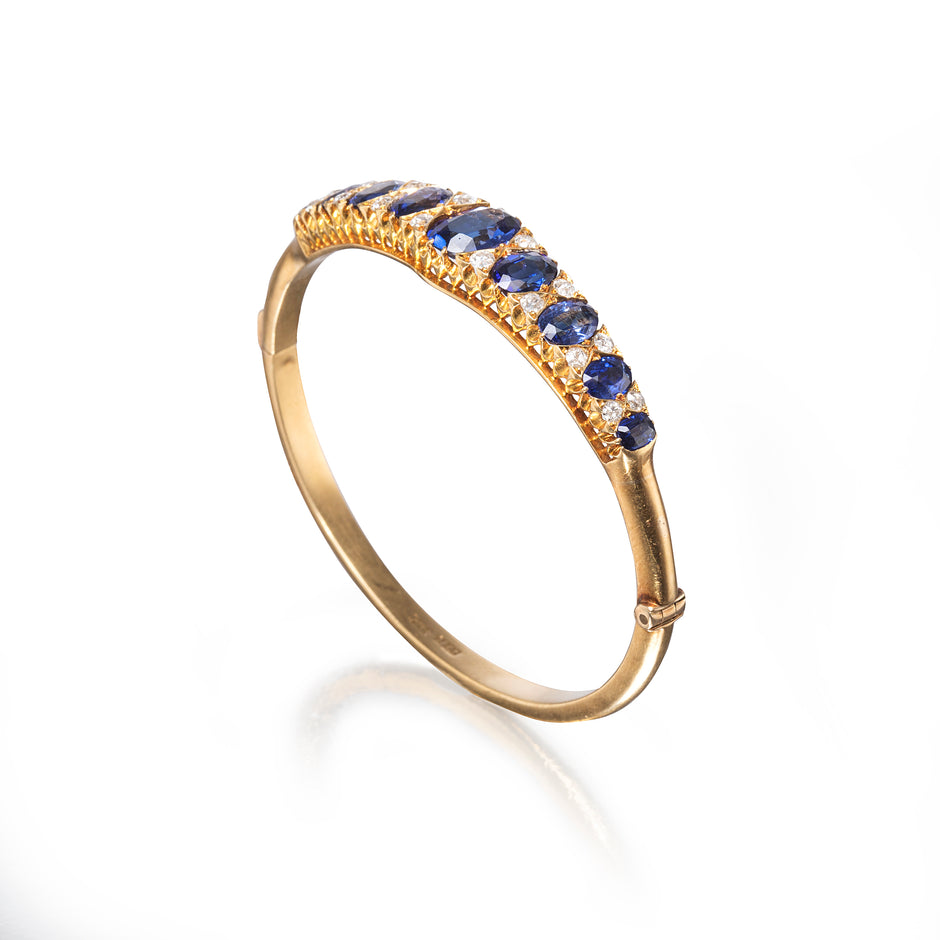 A gold Bangle featuring a curved row of round blue sapphires accented by small clear stones, shown on a clean white background with a soft reflection.