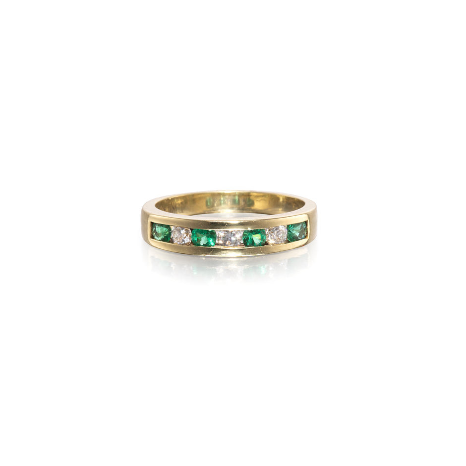 A yellow gold eternity ring featuring a channel-set row of round-cut green emeralds and clear diamonds, shown on a clean white background with a soft reflection.