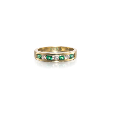 A yellow gold eternity ring featuring a channel-set row of round-cut green emeralds and clear diamonds, shown on a clean white background with a soft reflection.