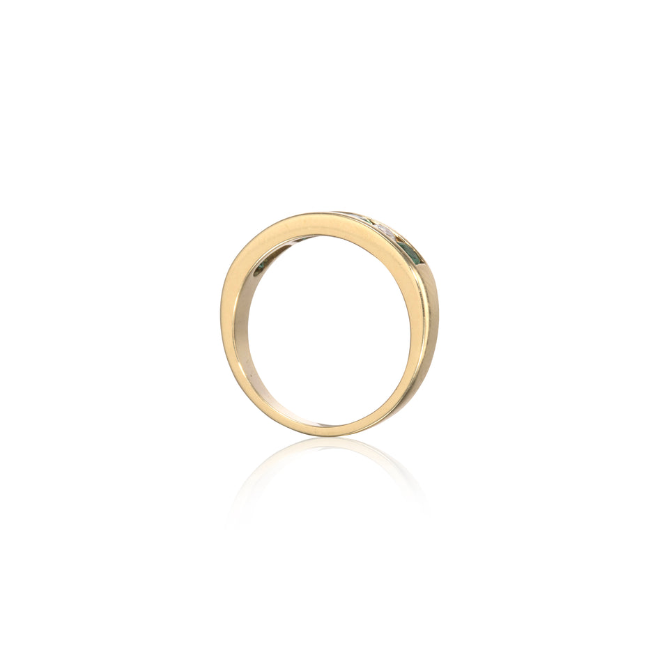 A polished yellow gold band ring with a small, clear, round-cut gemstone set on the upper right side, shown against a clean white background with a soft reflection below.