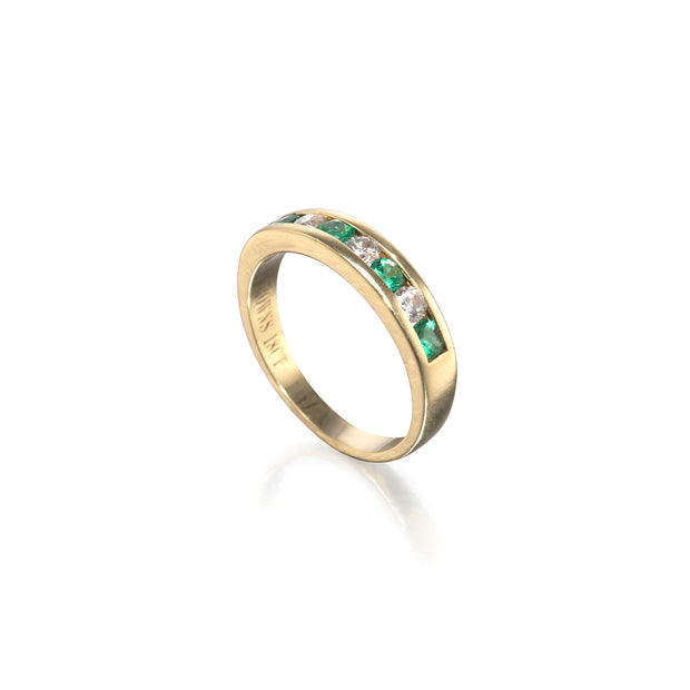 A yellow gold eternity ring featuring a channel-set row of green emerald-cut diamonds and clear round-cut diamonds, shown on a clean white background with a soft reflection.