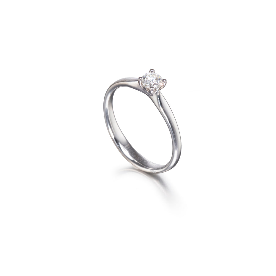 A platinum solitaire engagement ring with a round-cut diamond set in a four-prong setting, shown on a clean white background with a soft reflection.