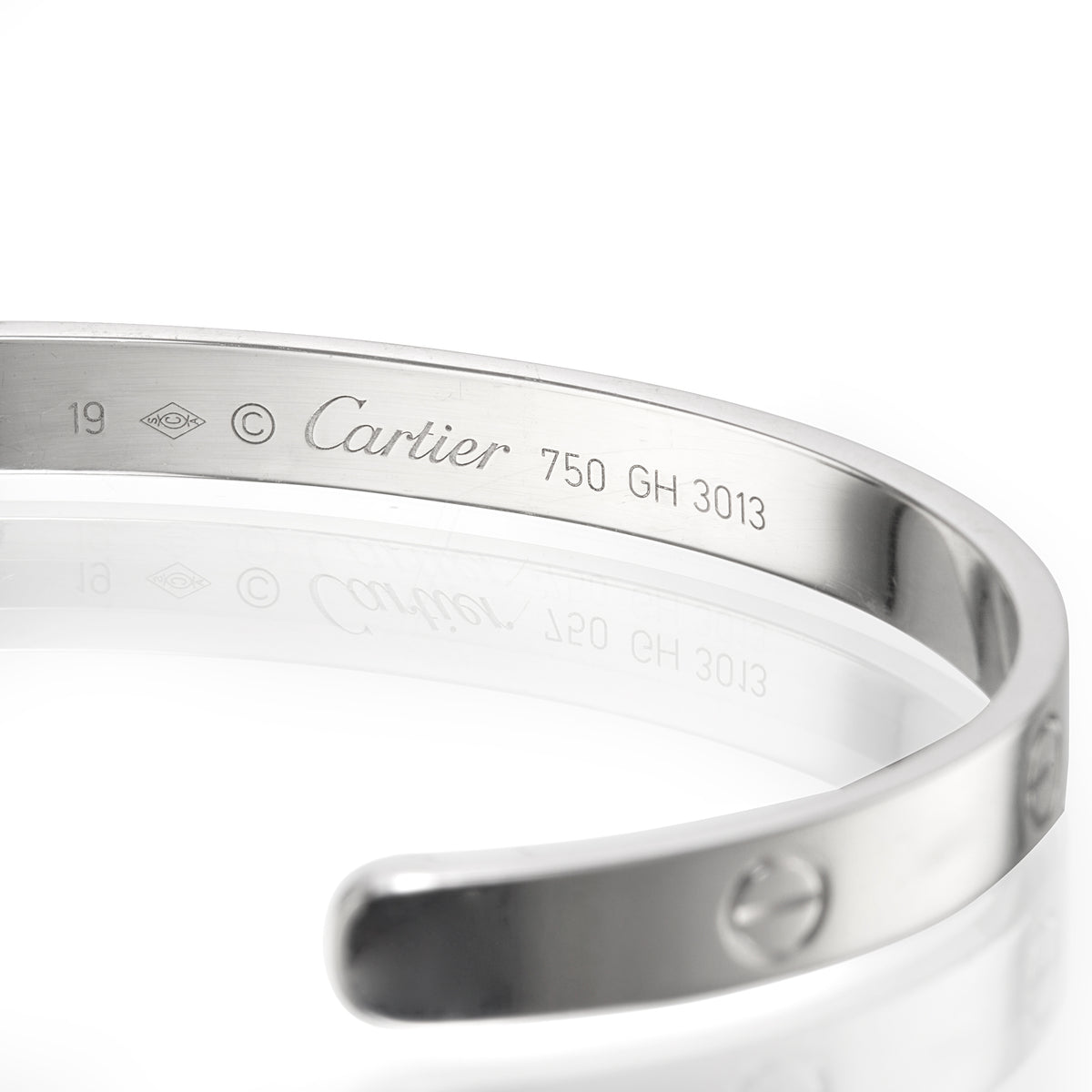 A close-up of a polished white gold Cartier bangle bracelet, showing the engraved brand name, 'Cartier', model number '750', and screw details on a reflective white surface.