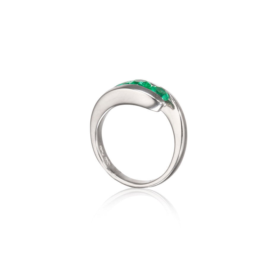 A platinum ring featuring a curved top set with three small vibrant green gemstones, shown on a clean white background with a soft reflection below.