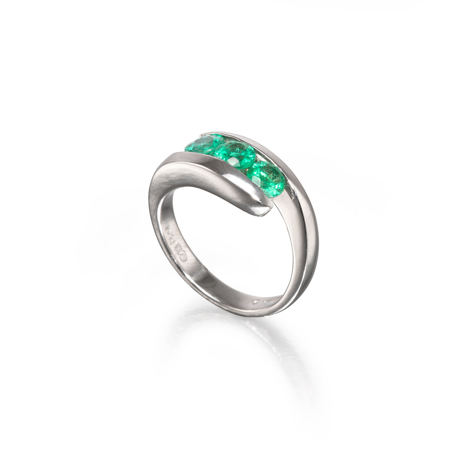A platinum curved ring featuring a row of vibrant green emerald-cut gemstones across the top, displayed on a clean white background with a soft reflection.