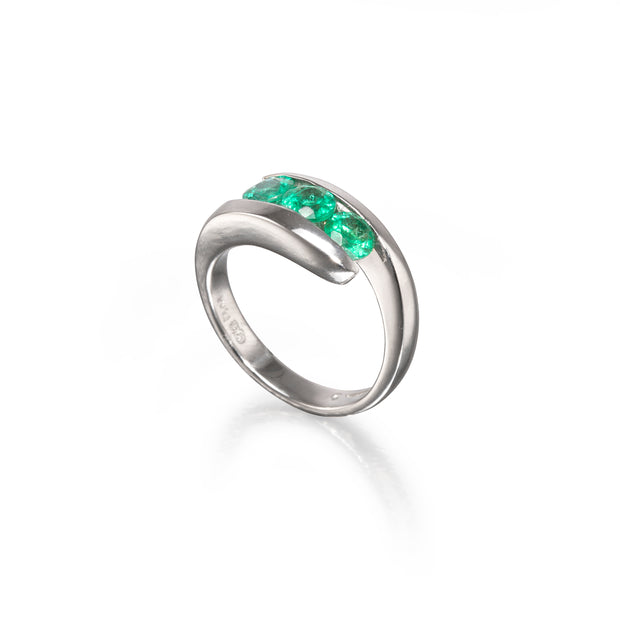 A platinum curved ring featuring a row of vibrant green emerald-cut gemstones across the top, displayed on a clean white background with a soft reflection.