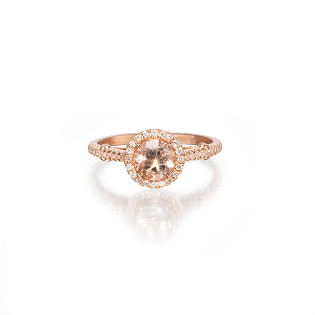 A rose gold halo ring featuring a round-cut peach morganite center stone surrounded by a halo of small clear stones, set on a white reflective background.