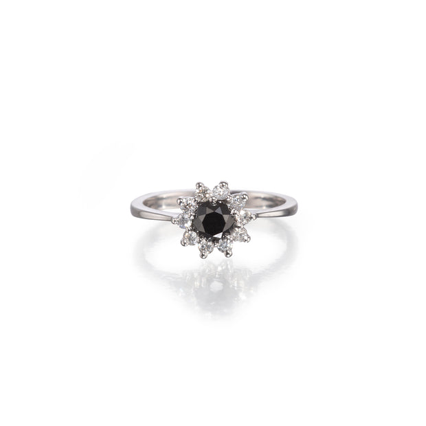 A white gold ring featuring a central round black gemstone surrounded by a halo of small clear stones, set against a clean white background with a soft reflection.
