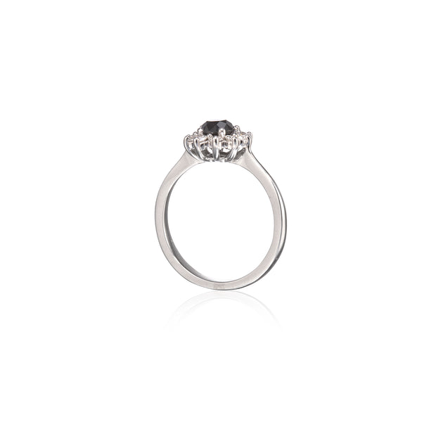 A white gold ring with a three-stone cluster setting featuring a dark round center stone and smaller clear side stones, shown on a plain white background with a soft reflection below.