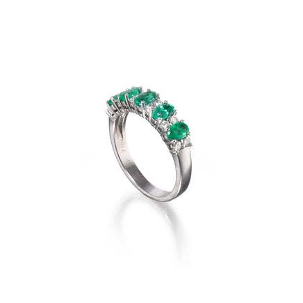A white gold ring featuring a row of oval-cut green emeralds and small clear accent stones, shown on a clean white background with a soft reflection.