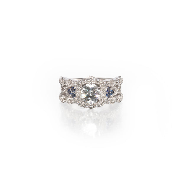 A white gold vintage-style ring featuring a central round-cut clear gemstone surrounded by a halo of smaller clear stones, with two small blue gemstones on each side, set against a plain white background.