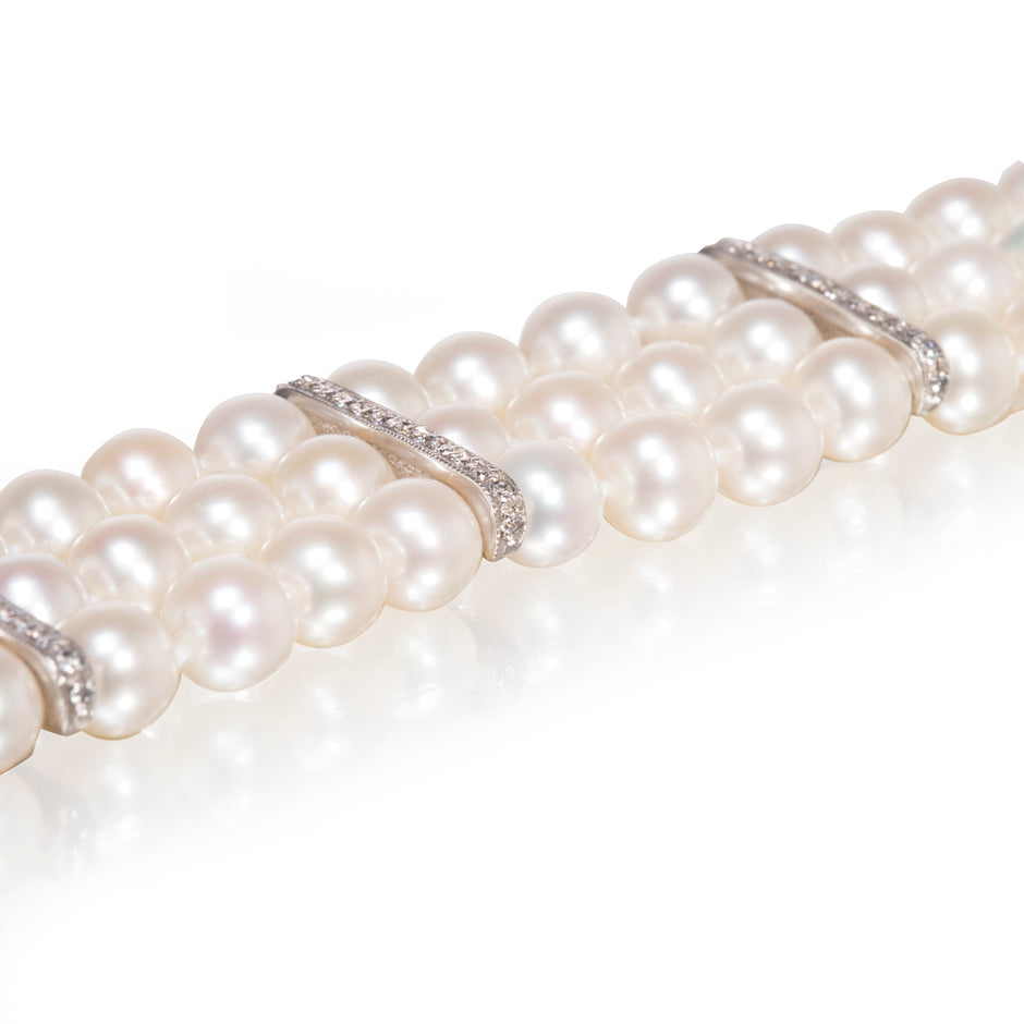 A close-up of a pearl strand bracelet featuring round white pearls and white gold settings with small clear stones, shown on a reflective white surface.