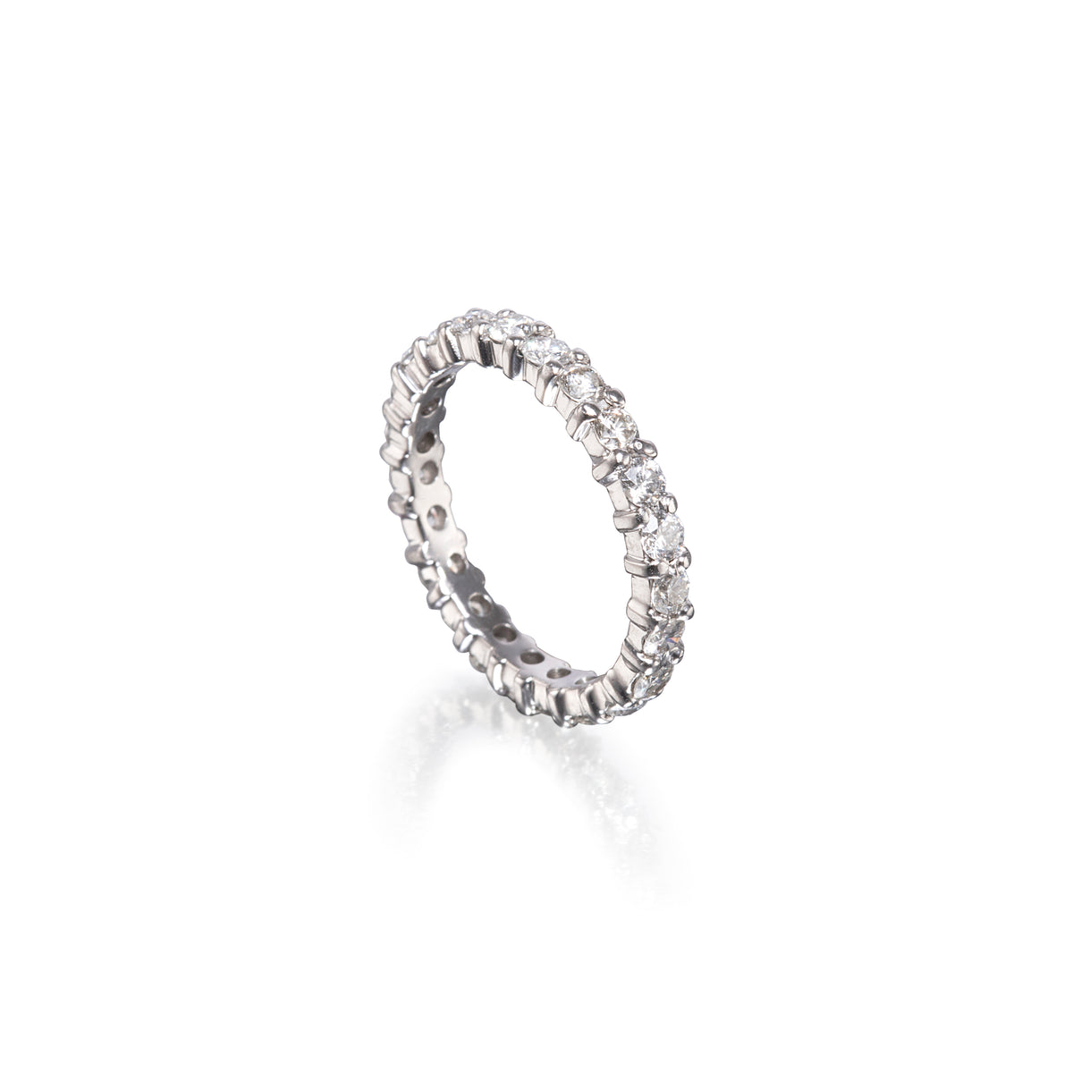 A platinum eternity ring set with small clear round-cut stones, shown at an angle on a clean white background with a soft reflection below.