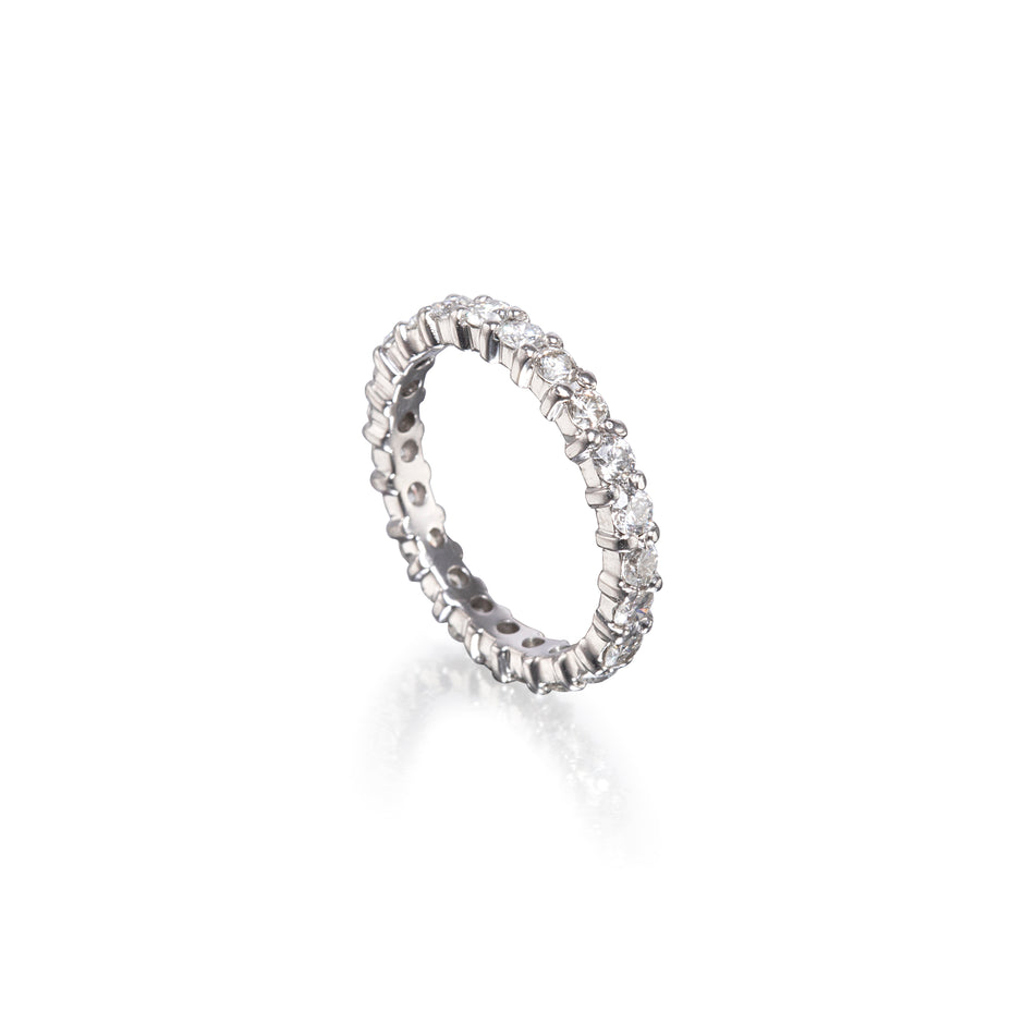 A platinum eternity ring set with small clear round-cut stones, shown at an angle on a clean white background with a soft reflection below.