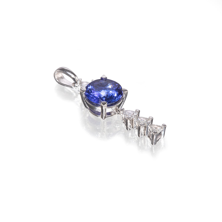 A platinum pendant featuring a round blue tanzanite center stone and three marquise-cut clear stones in a drop design, set on a white background with a soft reflection.