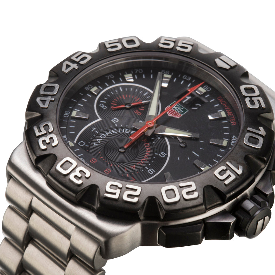 A close-up shot of a men's analog watch TAG, featuring a black dial with silver-tone hands, a red second hand, and a black bezel with white numerals, paired with a stainless steel link bracelet.