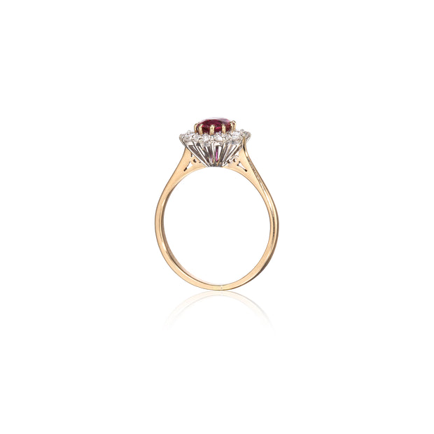 A yellow gold ring featuring a central round ruby gemstone surrounded by a halo of small clear stones, set against a plain white background with a soft reflection below.