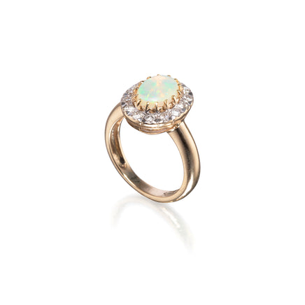 A yellow gold ring featuring a round cabochon opal in a halo setting with small clear stones, shown on a clean white background with a soft reflection.